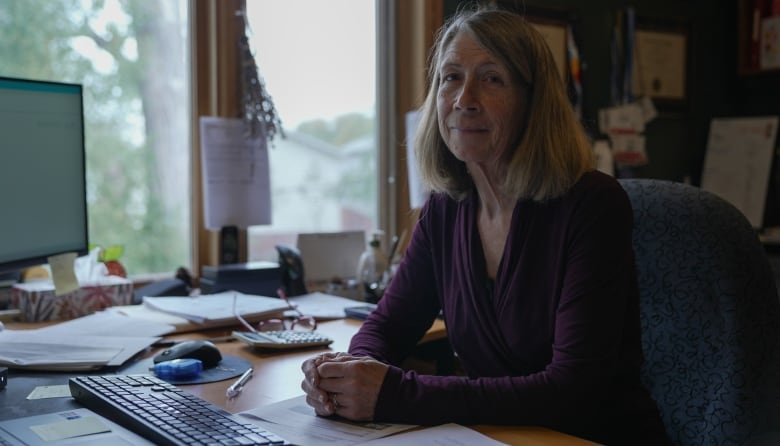 A woman sits at a desk, looking at the camera.