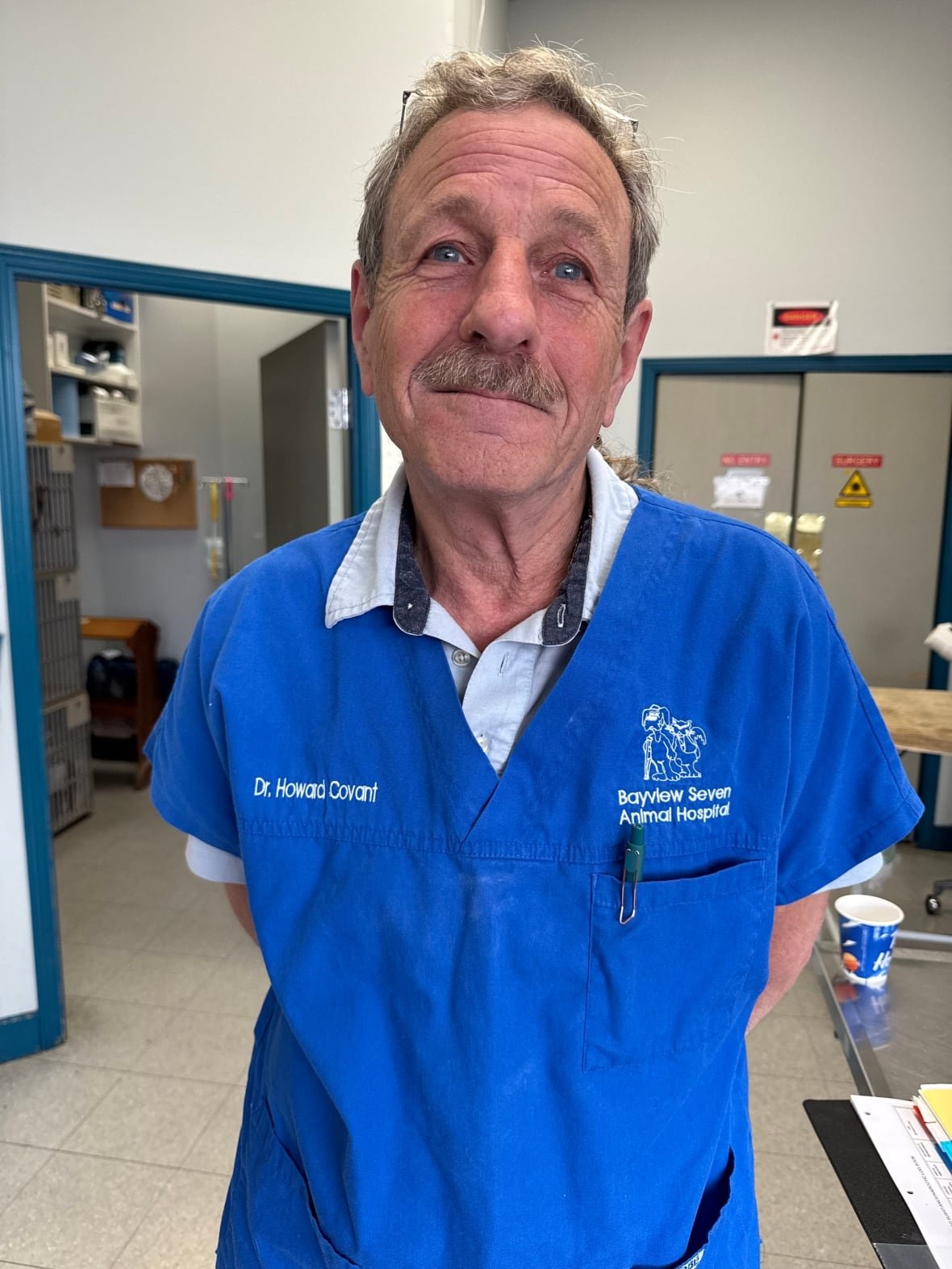 A veterinarian wearing a blue coat stands in his office looking at the camera