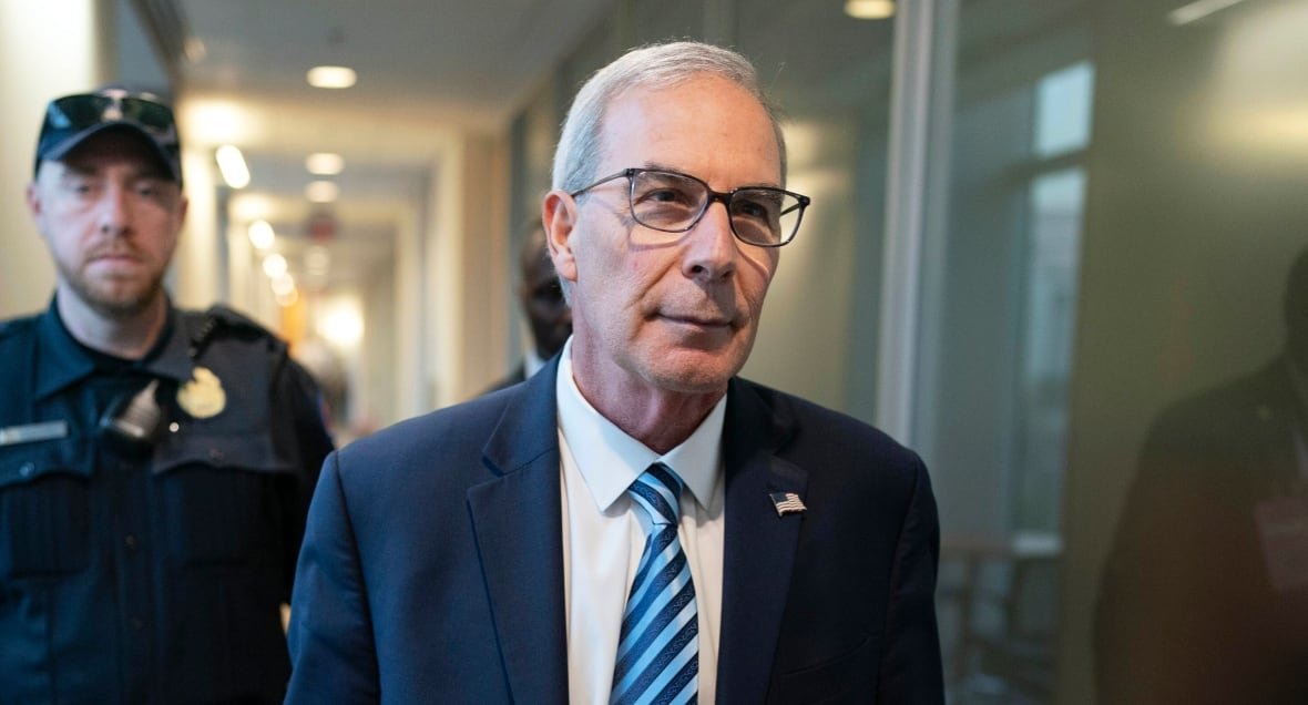 A man in a navy blue suit with a light blue striped tie carries a briefcase as he walks through a building. His hair is grey and he is wearing black glasses. He has an American flag pin on his lapel.