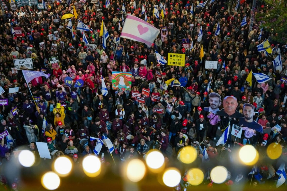 Hundreds of demonstrators wave Israeli flags and signs during a protest. One yellow sign says "BRING THEM HOME".