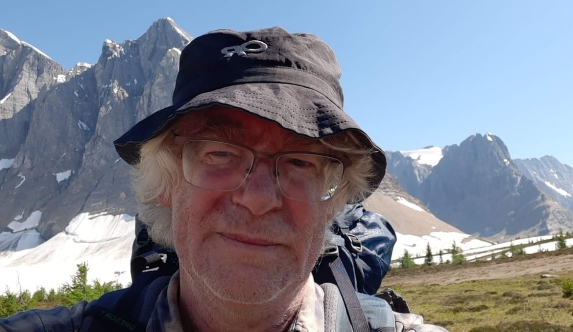 A man wearing a hat looks at the camera with a backdrop of mountains pictured behind him.