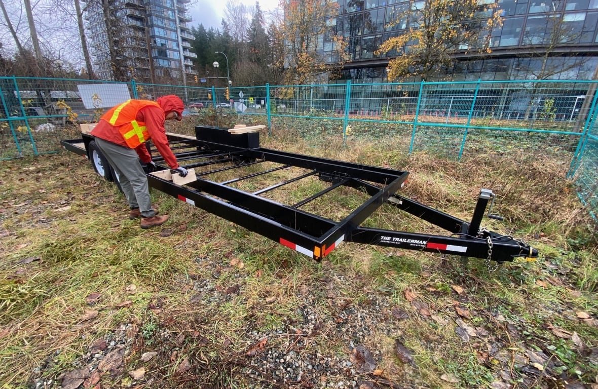 In a grassy area fenced off by green fences, a person in an orange and yellow visibility vest bends over a trailer hitch. Skyscrapers and other buildings are visible beyond the fences.