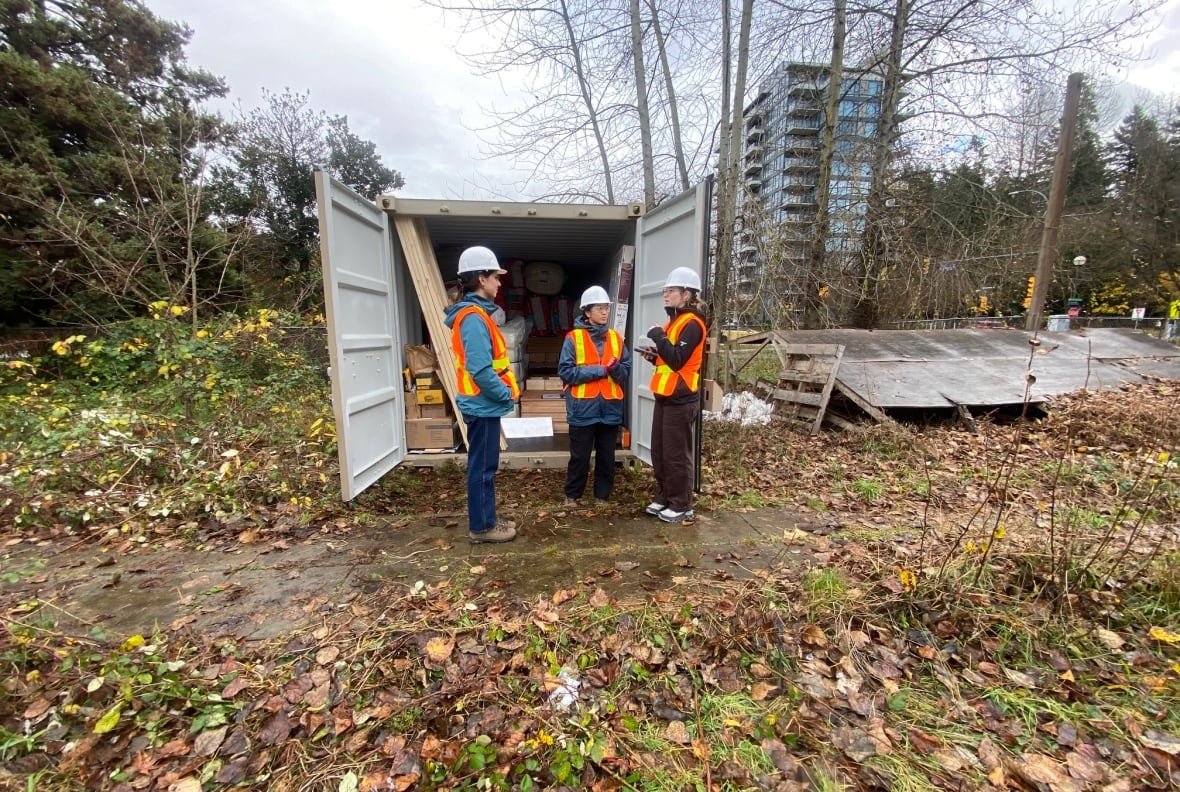 Three people in orange and yellow visibility vests and hard-hats stand outside the open doors of what looks like a shipping container, talking to each other. They are outside on a dirt path with fallen leaves around them, and a city building visible in the back beyond the trees.