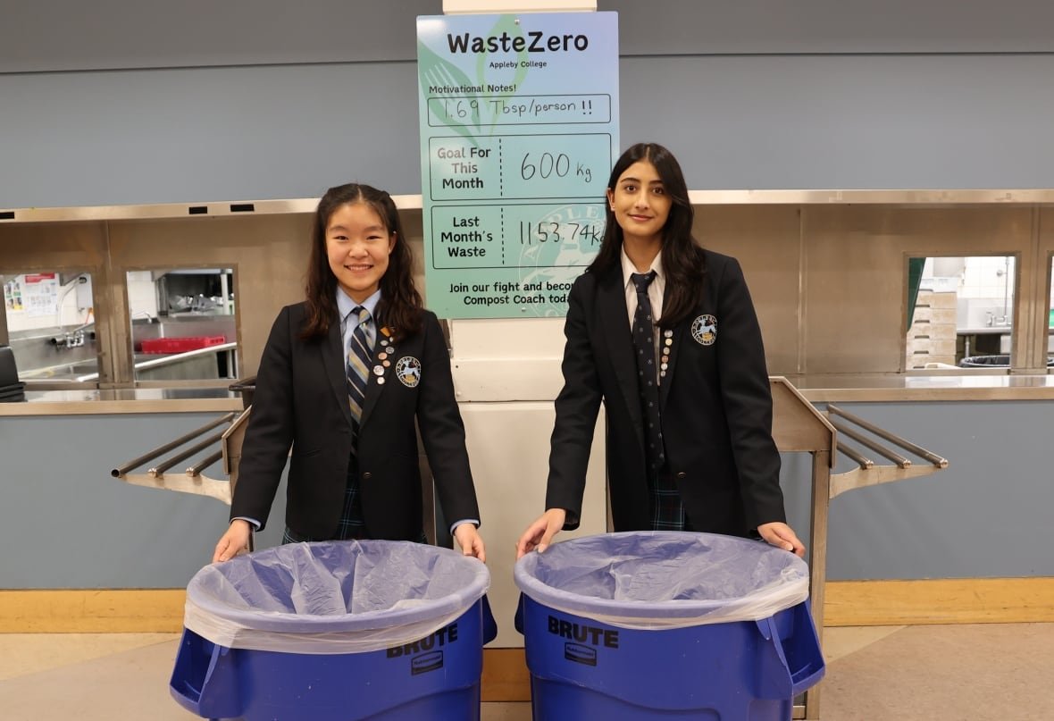 Two girls in uniforms in front of the serving counter of a cafeteria with blue trash bins in front of them