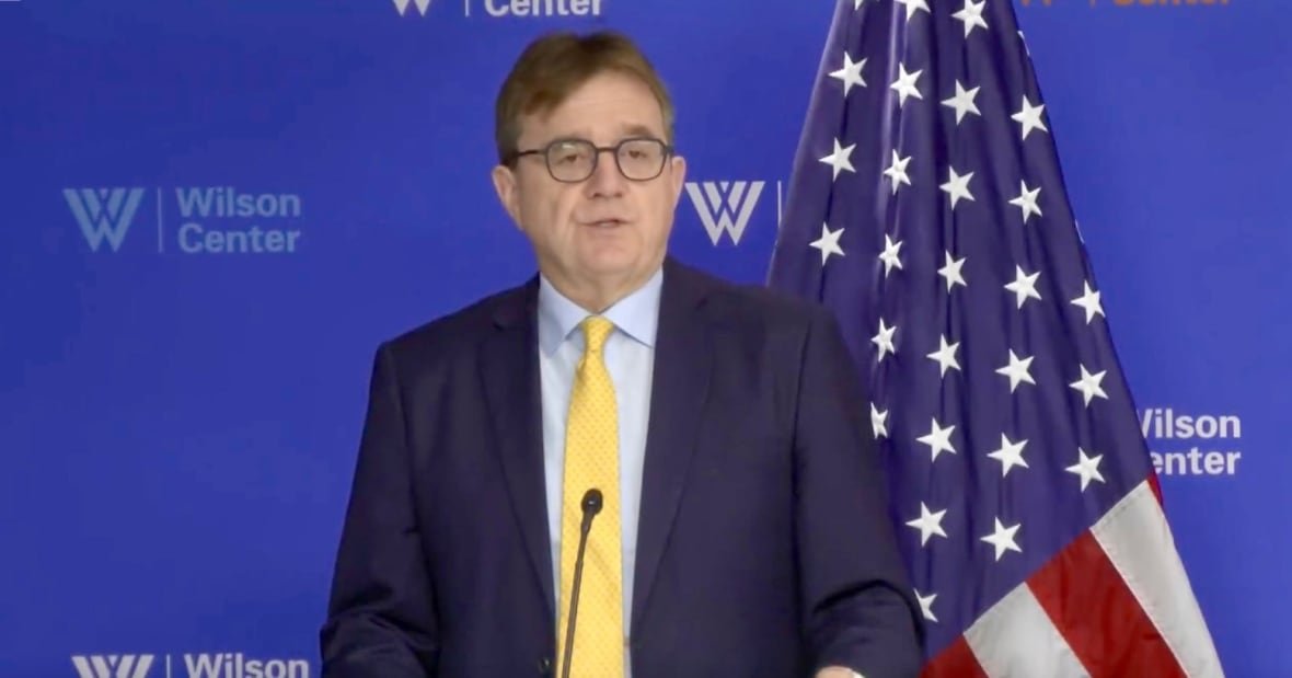 A man in a suit speaks in front of a U.S. flag.