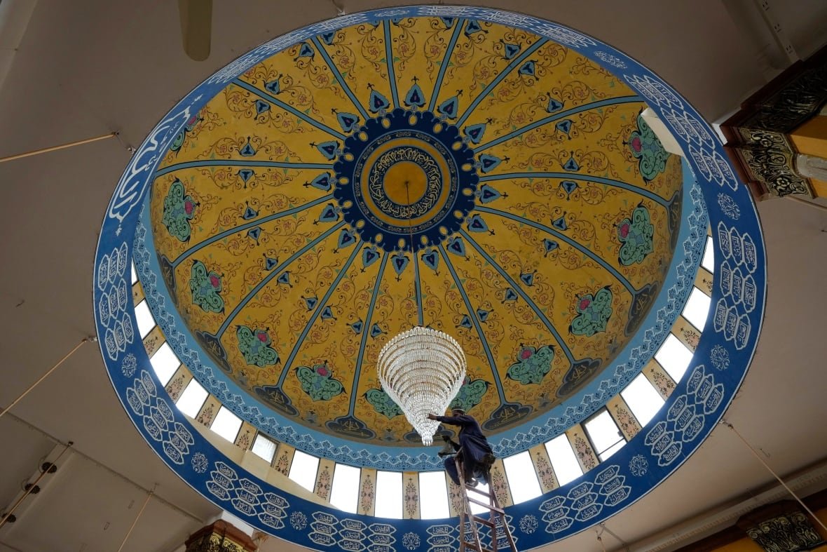 Viewed from the ground, a worker on a ladder cleans a chandelier inside a mosque