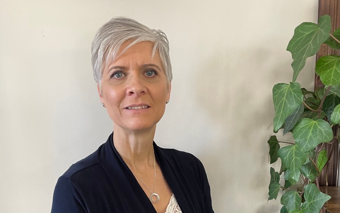 A woman with short silver hair stands indoors next to a long, leafy houseplant.