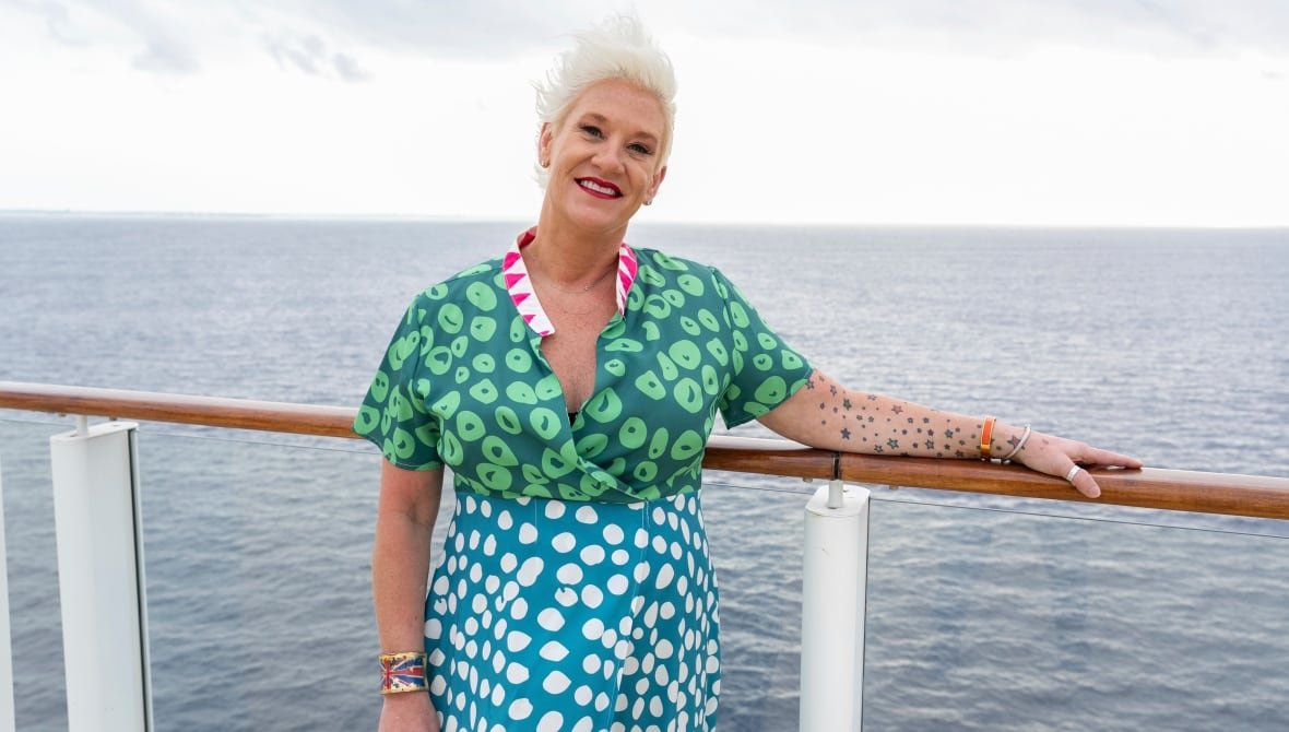 2024-chefs-making-waves A woman white short, spiky platinum blonde hair poses against the white railing of a boat in a colourful, short-sleeved dress.
