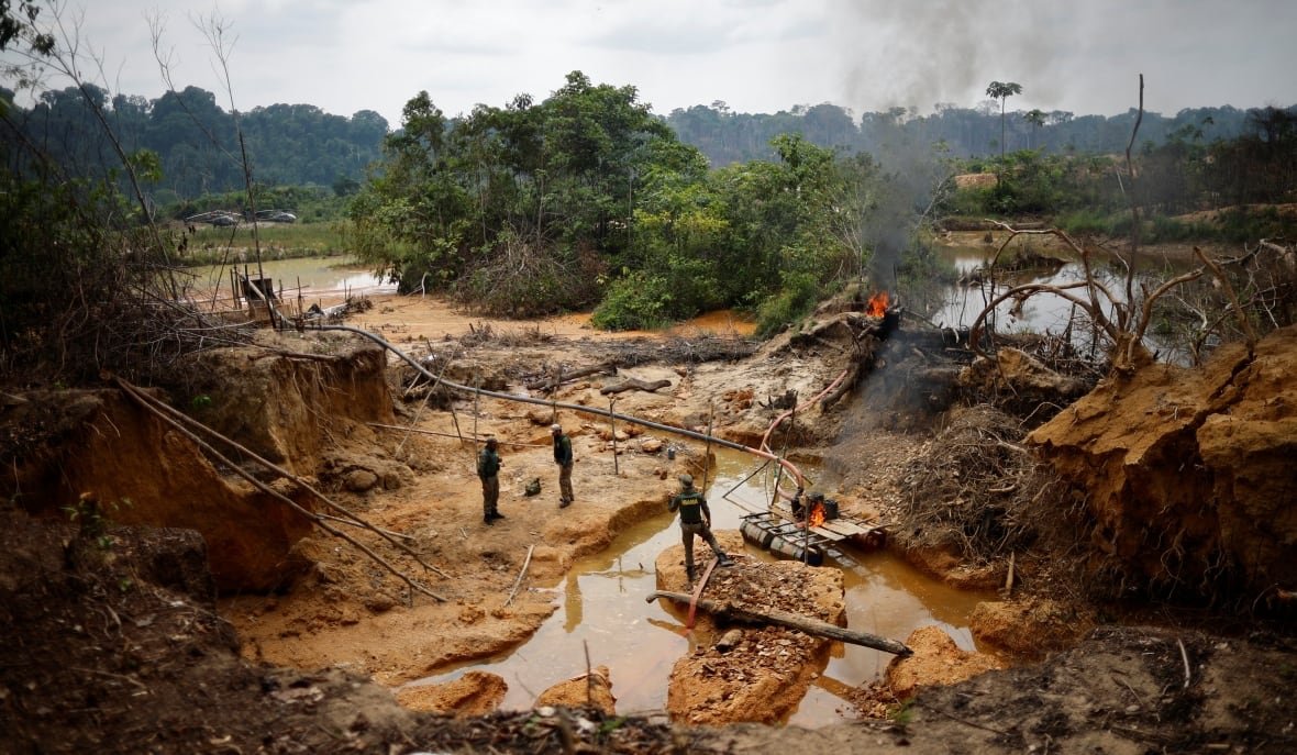 A muddy area is shown, with three people standing in the middle of it. Trees are visible past them, with a river behind it.