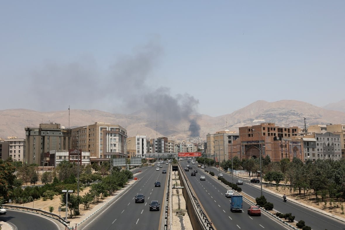 iran-nuclear-opposition-moment-1 Smoke rises from a city in front of a highway, with mountains in the background.
