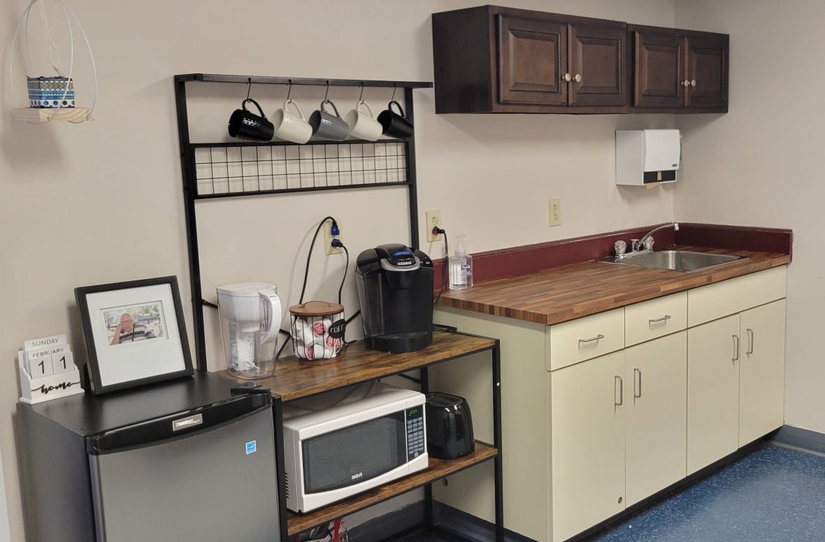 kitchenette-palliative-care-room A countertop with a sink and cabinets above it. At the side, there is a small cart holding a Keurig and microwave with a mini fridge next to it.