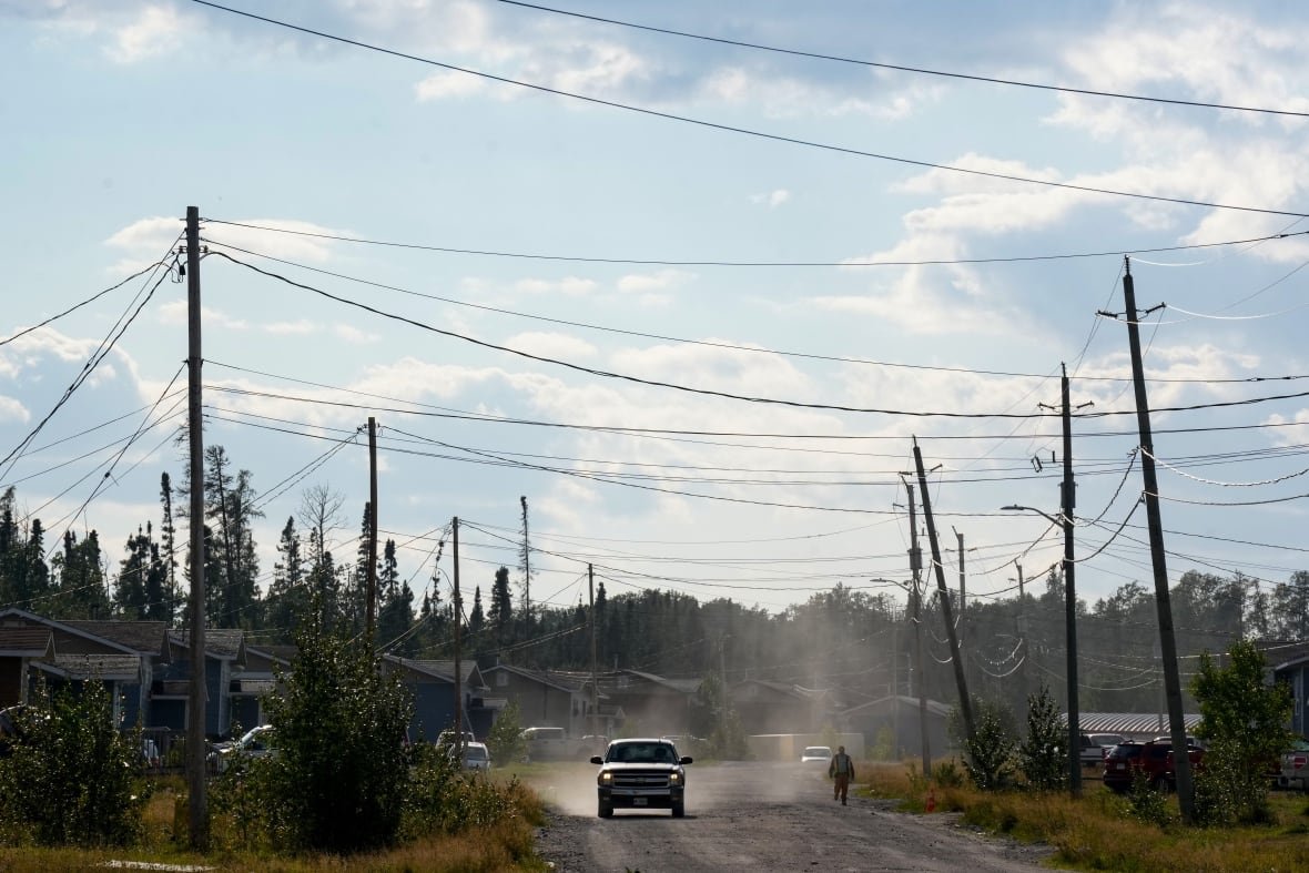 ont-mining-20250417 A car drives through Neskantaga First Nation in Neskantaga, Ont., on a dirt road with houses along the side and teleophone wires overhead on a sunny summer day.