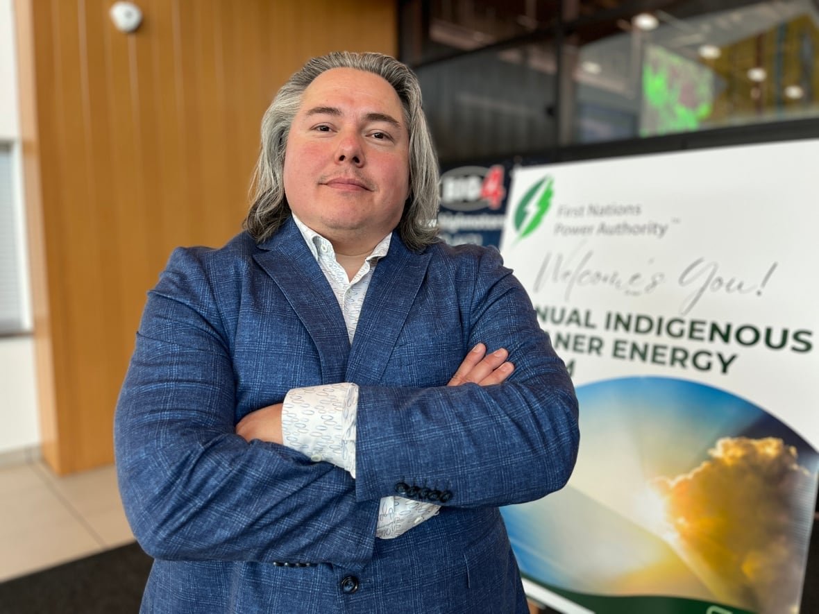 steven-vavaida-1 A man with long hair, a blue blazer and a dress shirt is pictured with his arms crossed in front of a conference sign.