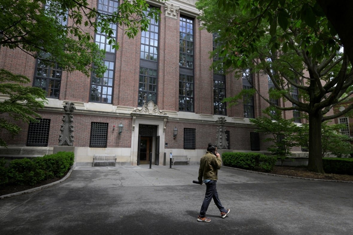usa-trump-harvard A student in jeans and a green jacket walks on a university campus with a brick building.