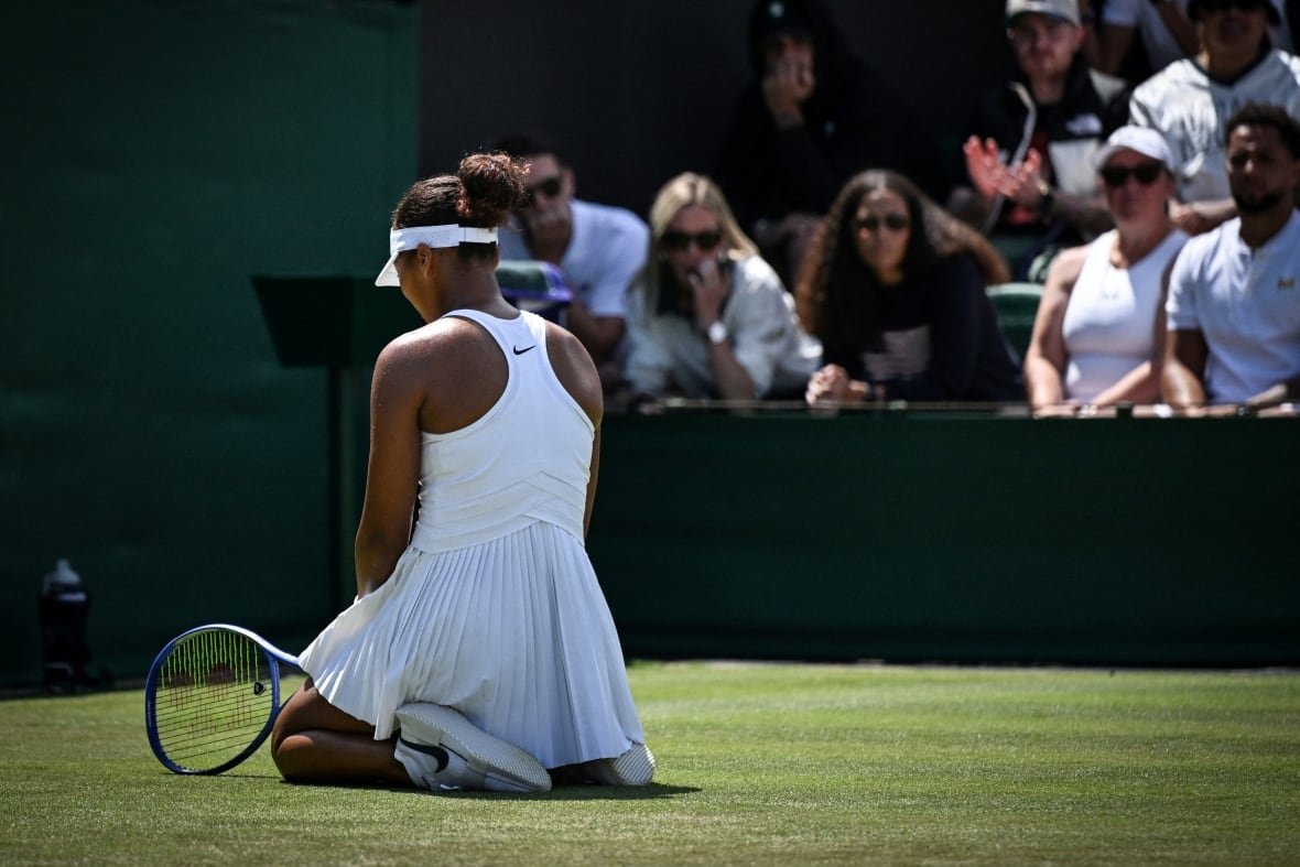2222779593 A women's tennis player, with her back to the camera, kneels on the grass court with her racquet touncing the ground.