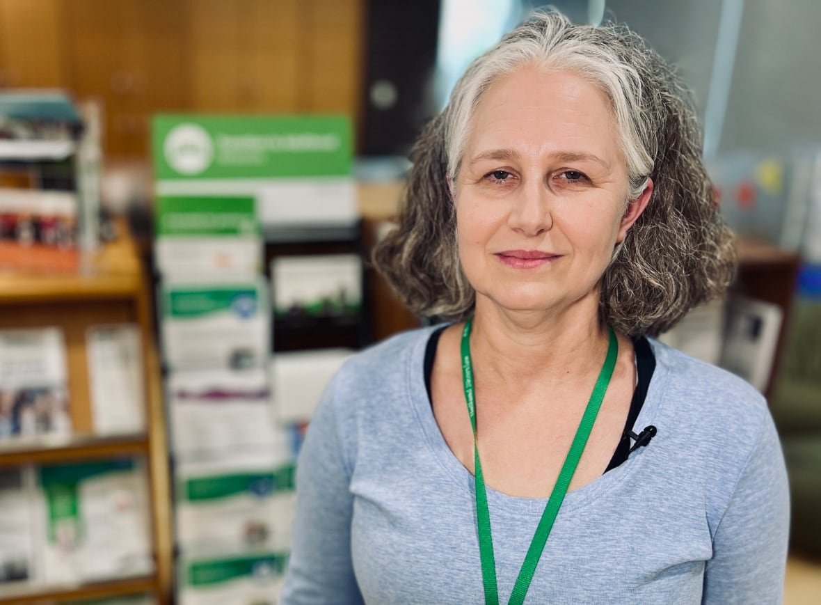carolyn-mcdougall A woman with wavy, shoulder-length hair and wearing a pale blue long sleeved top smiles slightly while standing in an indoor room, with shelves of pamphlets behind her.