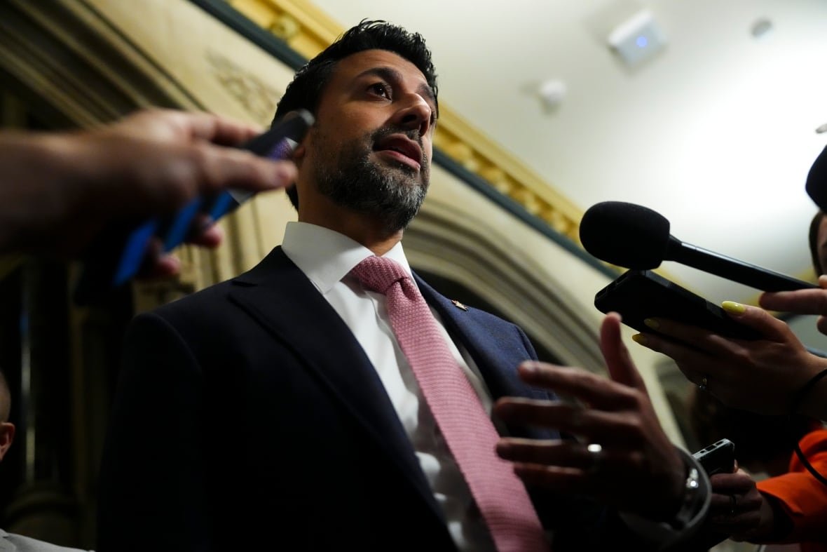 caucus-20250611 A man with dark skin and short black hair speaks into microphone. He's wearing a blue suit and pink tie