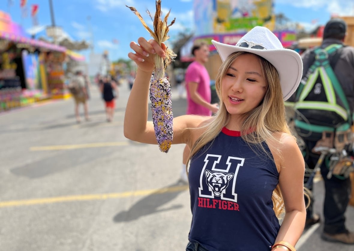 cgy-stampede-2025-1 A woman in a t-shirt and cowboy hat holds up a cob of toasted street corn.