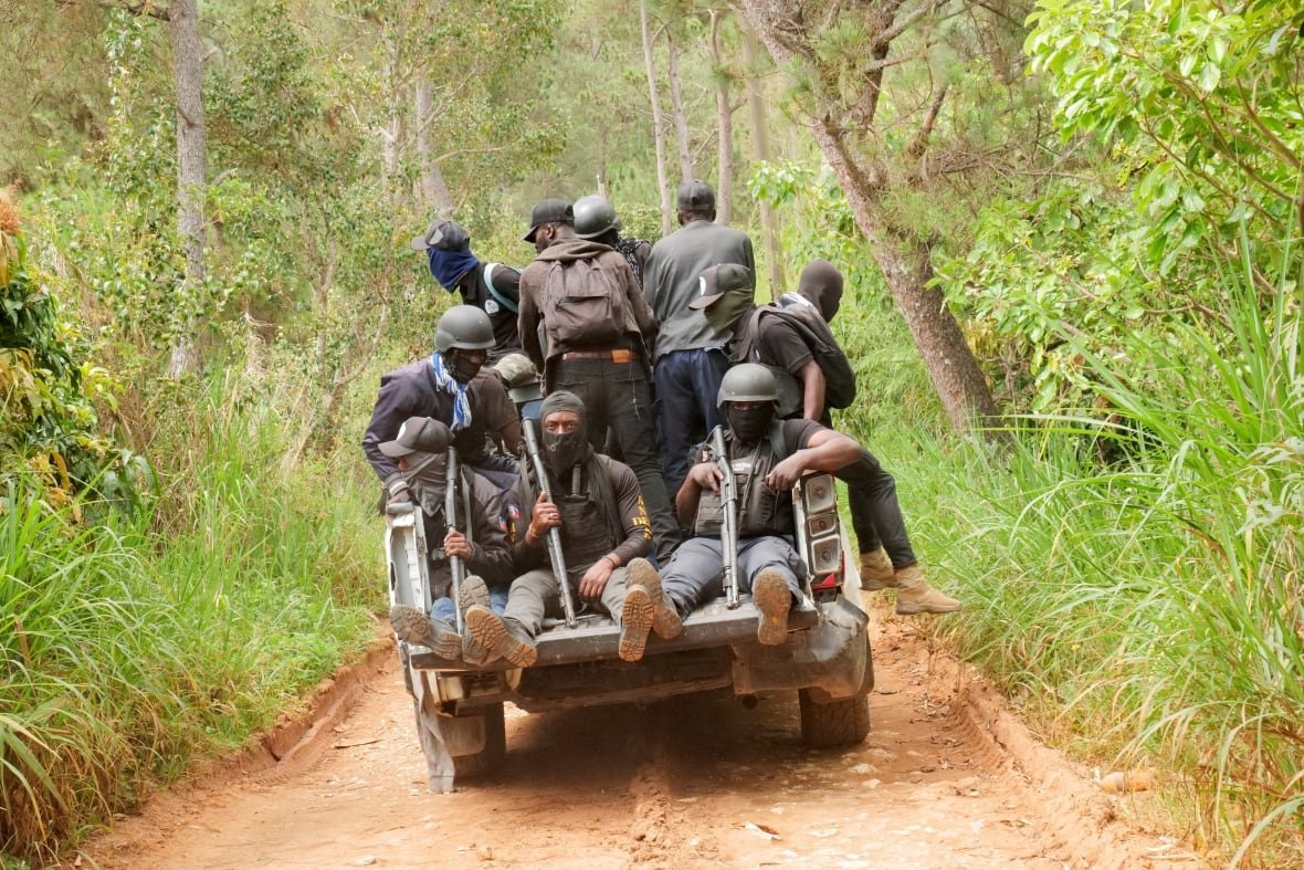 Several men with masks and hoods and carrying weapons are shown travelling in the back of a pickup truck down a rural road.