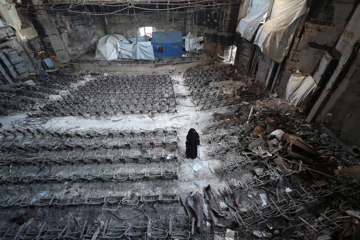 israel-palestinians-gaza-displaced A woman in a black hijab stands on steps between rows of damaged seats in a lecture hall inside a ruined university building.