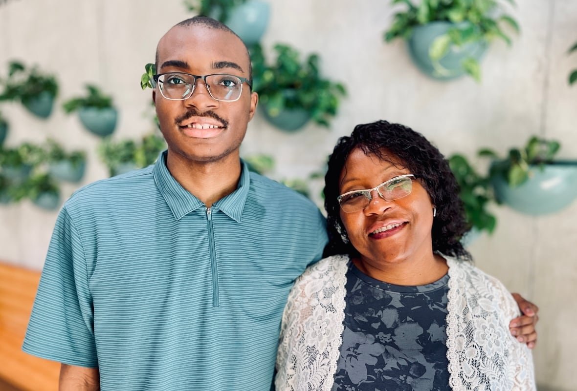 jordan-o-neal-and-brendora-paul A smiling young man in glasses and a teal polo shirt stands with one arm around a smiling shorter woman wearing a blue floral-patterned dress with a white lace jacket.