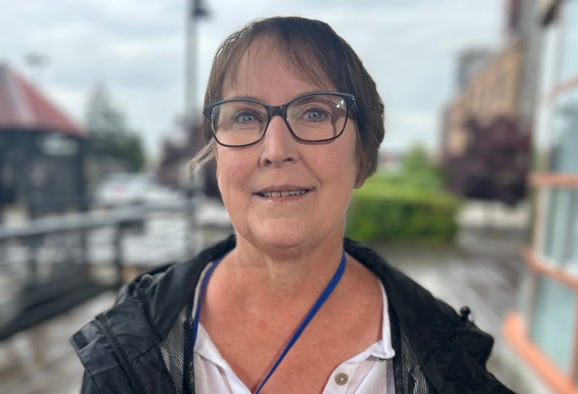 rachelle-hole A smiling brunette woman in glasses, a white top and black wind jacket stands outside under a building overhang on a grey, rainy day.