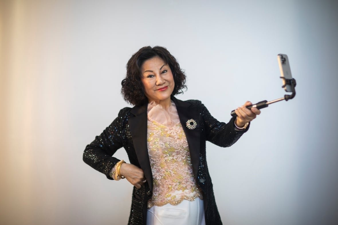 An East Asian woman with a dark bob haircut smiles while holding a selfie stick. She wears a sparkly pink and green top with a sparkly black blazer over top.