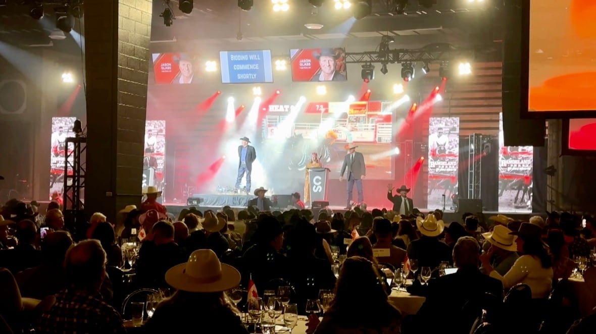 tarp-auction Bright white and red lights shine down on a stage as a man with a cowboy hat stands in front of a crowd.