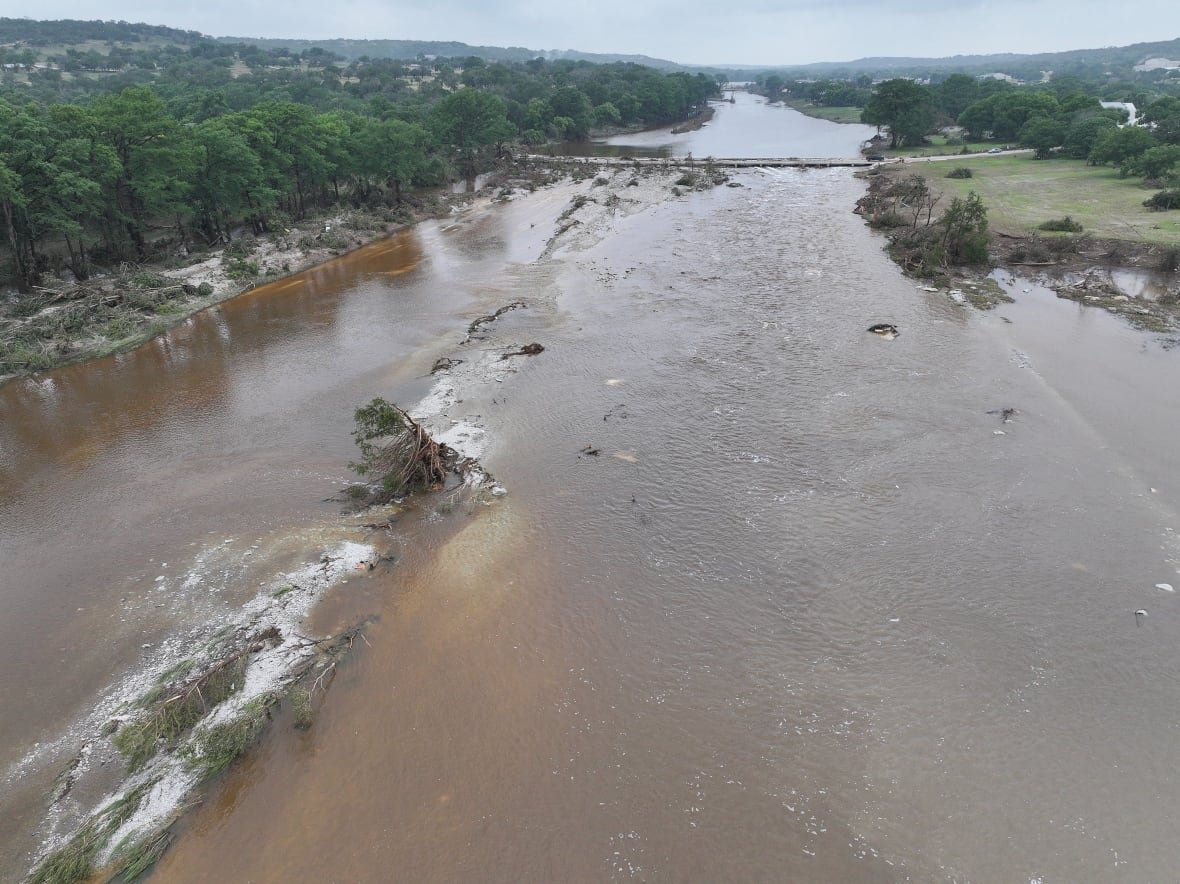 A swollen brown river is seen with trees on its banks.