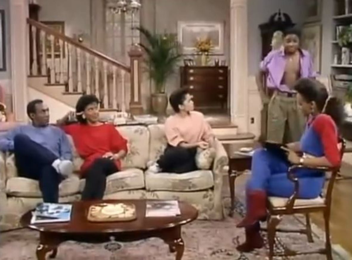the-cosby-show-theo-huxtable A still frame from an episode of The Cosby Show showing the inside of a home, with a large staircase in the background, with man, a woman and a young woman sitting on a couch, a young man standing to the right of the couch and to the left of a young woman sitting in a chair.