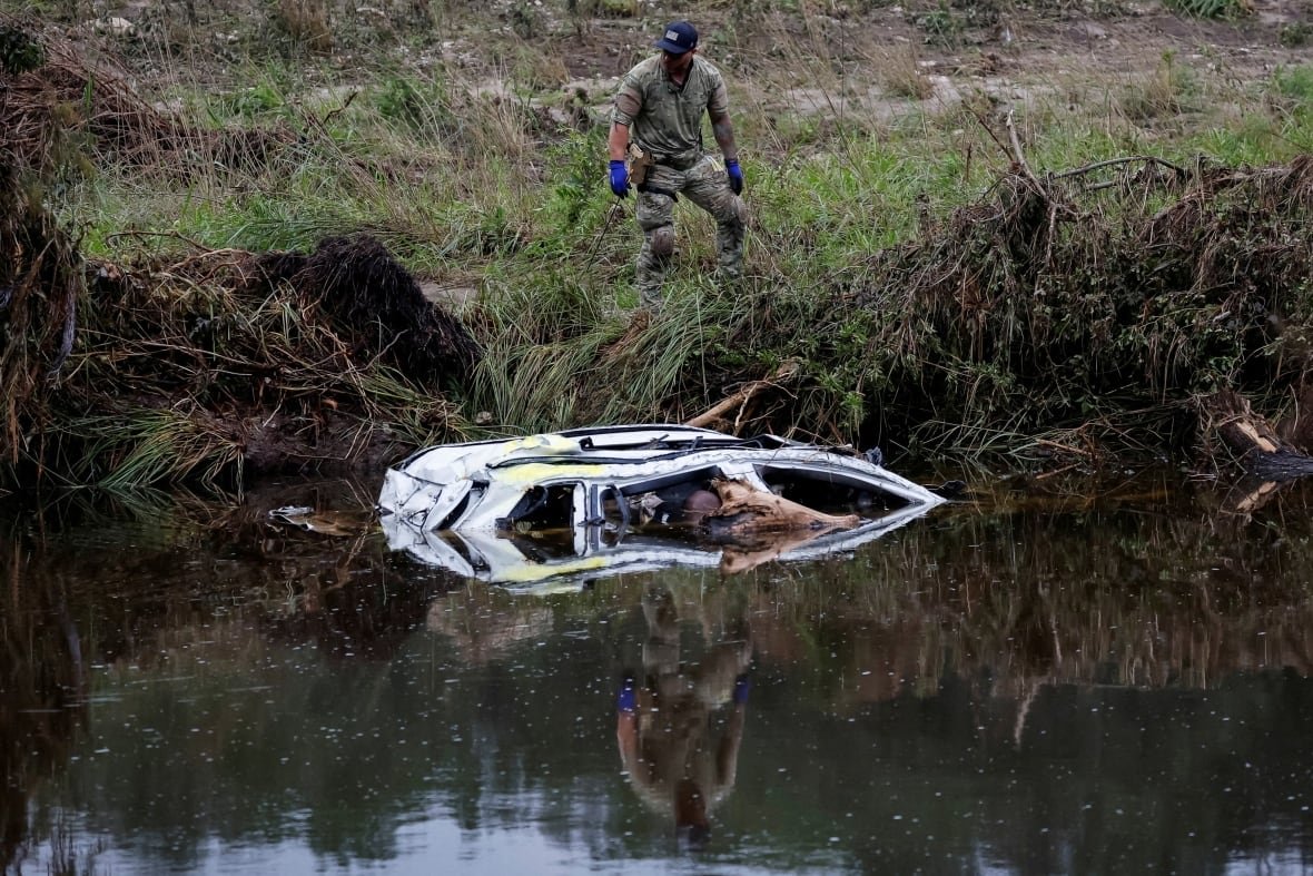 usa-weather-texas-flooding-4 A person on a river's bank surveys a car that's partially submerged in the war.