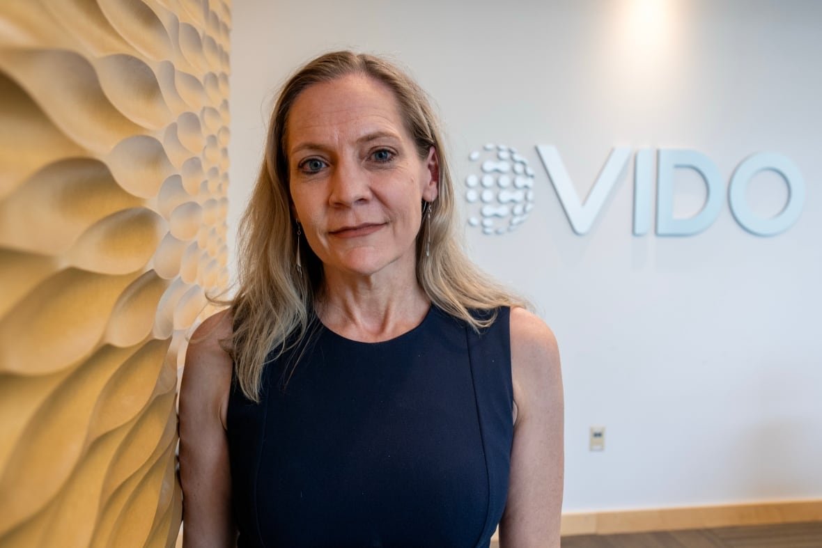 sask-vaccine-research-20250925-1 A blond woman in a black shirt stands against a textured wall with a sign that reads VIDO behind her.