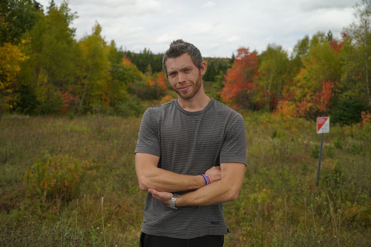Man in grey t-shirt stands near trees.