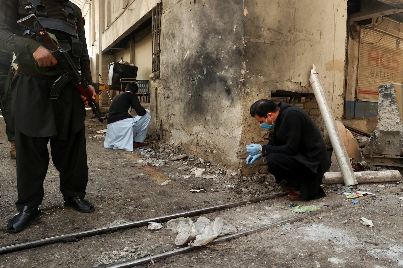 A man in a black robe, wearing gloves and a face mask, crouches on the ground near a building blackened with smoke.