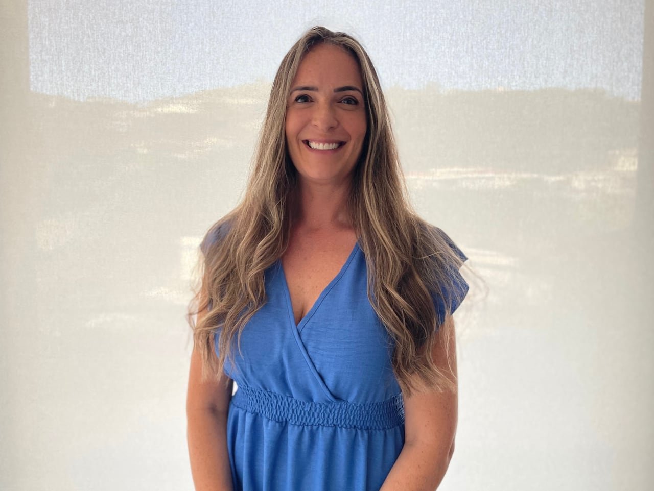 A woman with curly long brown hair in a blue dress with short sleeves stands in front a cream-colored wall outside, smiling at the camera.