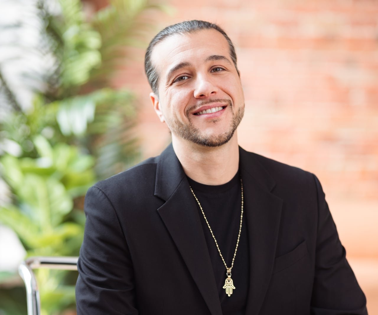 A man with short black hair and a short beard in a black suit and shirt sits on a chrome chair, smiling, with a brick wall and plant in the background.