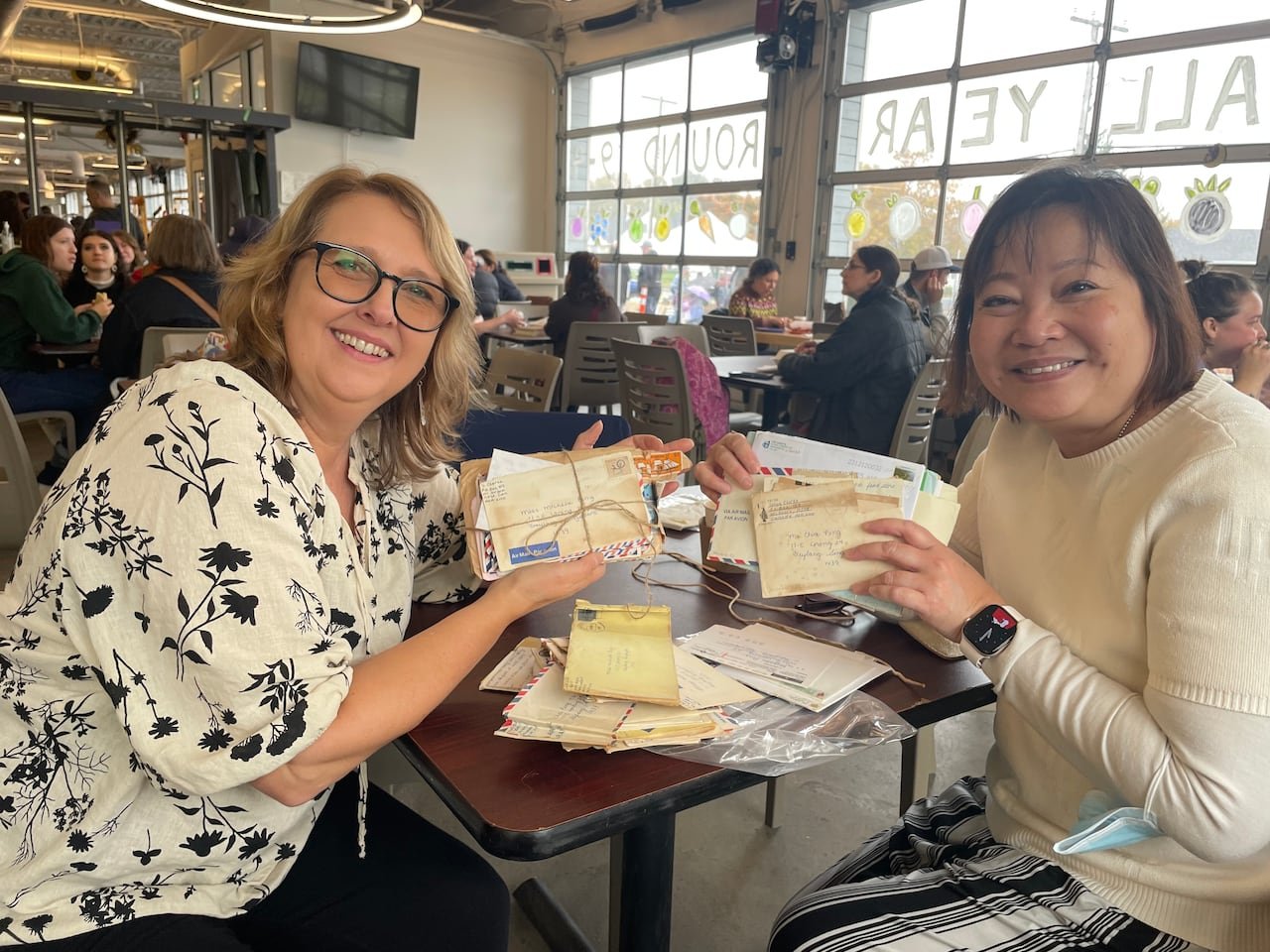 Two women sitting at a table in food court with stacks of envelopes and letters open