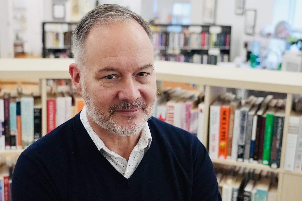 A man in a button-up shirt and navy sweater stands in a bright indoor space, rows of bookshelves full of books behind him.