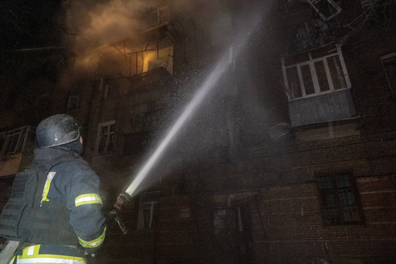 A emergency responder dressed in a uniform with light-reflective stripes and a helmet holds a large hose as it sprays water at a building that's on fire at night.