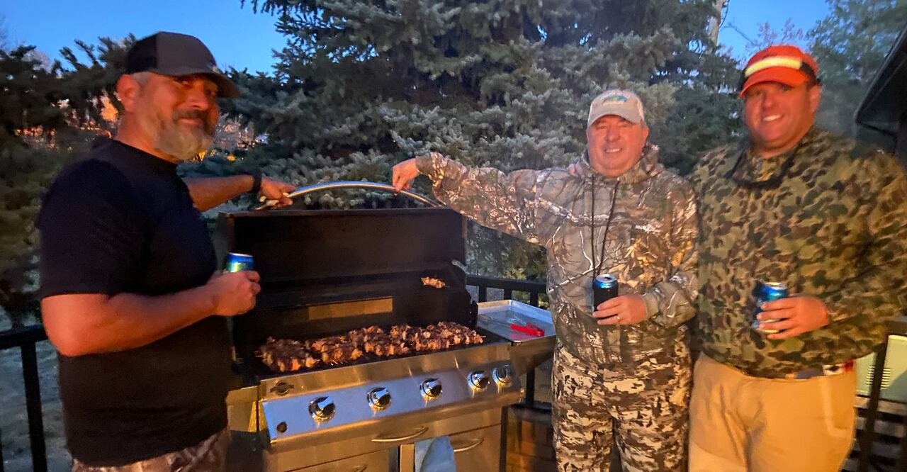 Three men in camouflage attire gather around a stainless steel barbecue grill cooking meat.