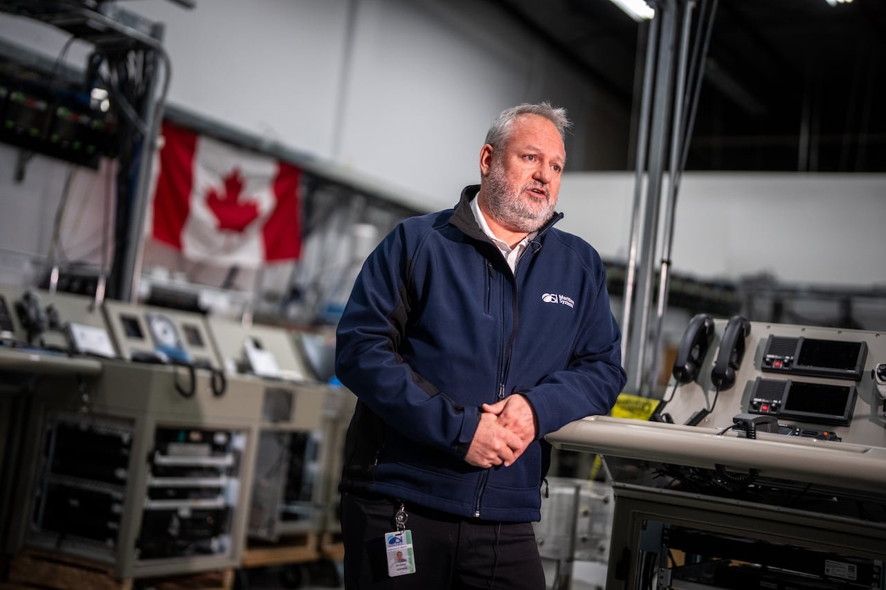 A man wearing a jacket stands inside a lab, with electronic consoles and a Canadian flag visible behind him.