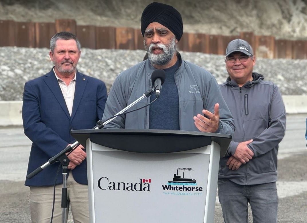A man stands speaking at a podium outdoors, with two other people looking on.