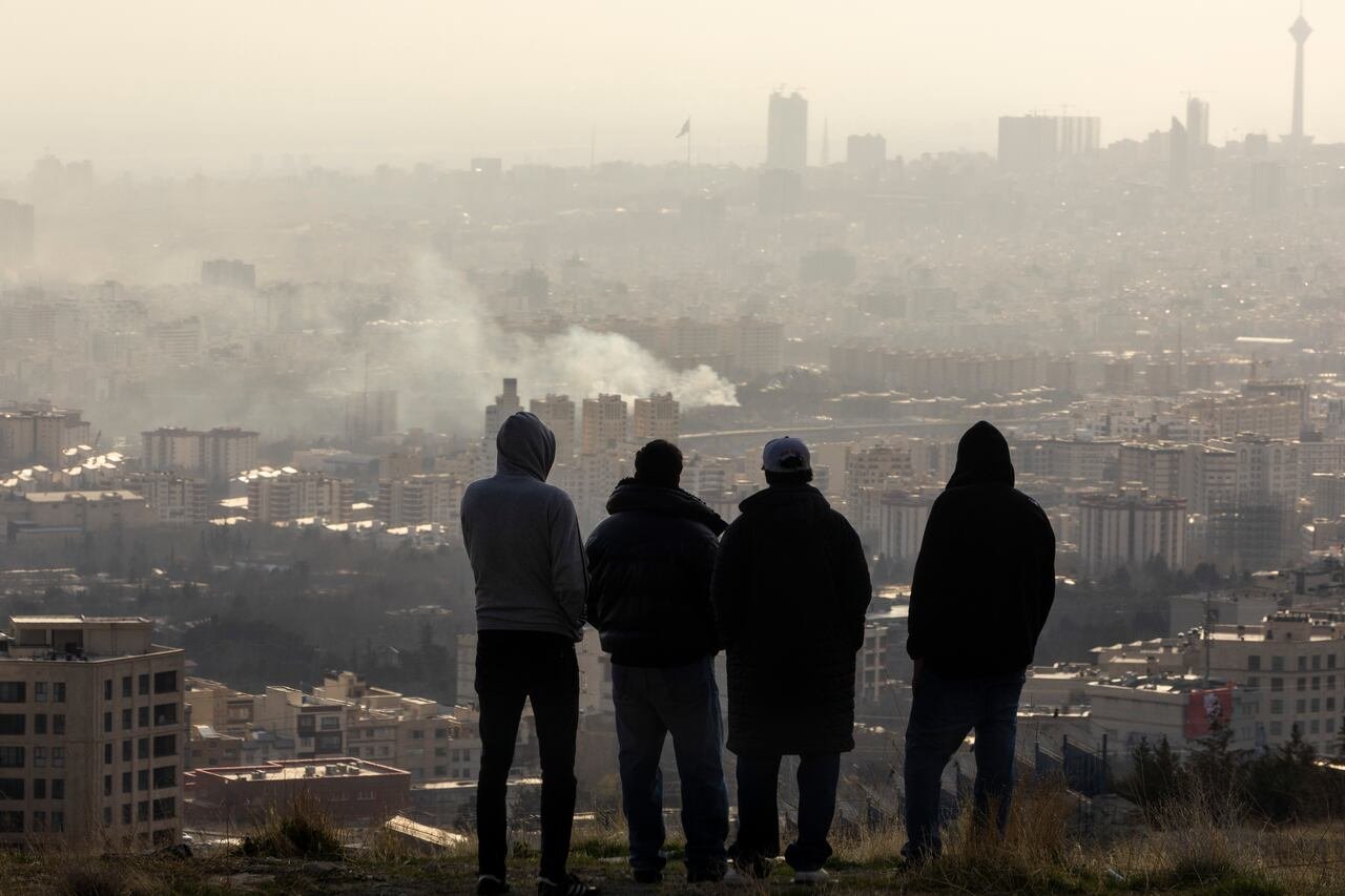 Young men silhouetted from behind as they stand on on a hill looking at smoke rising from the buildings of a city