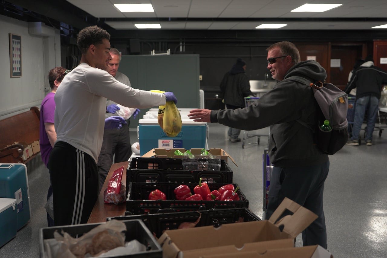 default-64 A young man hands a bag to an older man wearing sunglasses and a coat in what appears to be a transaction. The two people are separated by a table with fruits and vegetables on it.