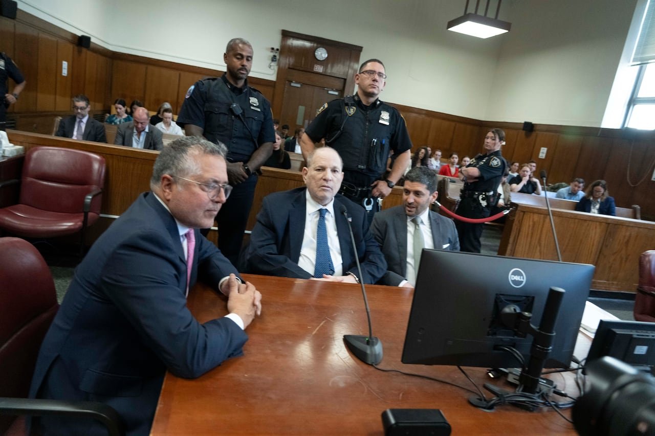 A man in a suit sits at a table in courtroom in front of two police officers and two men in suits.